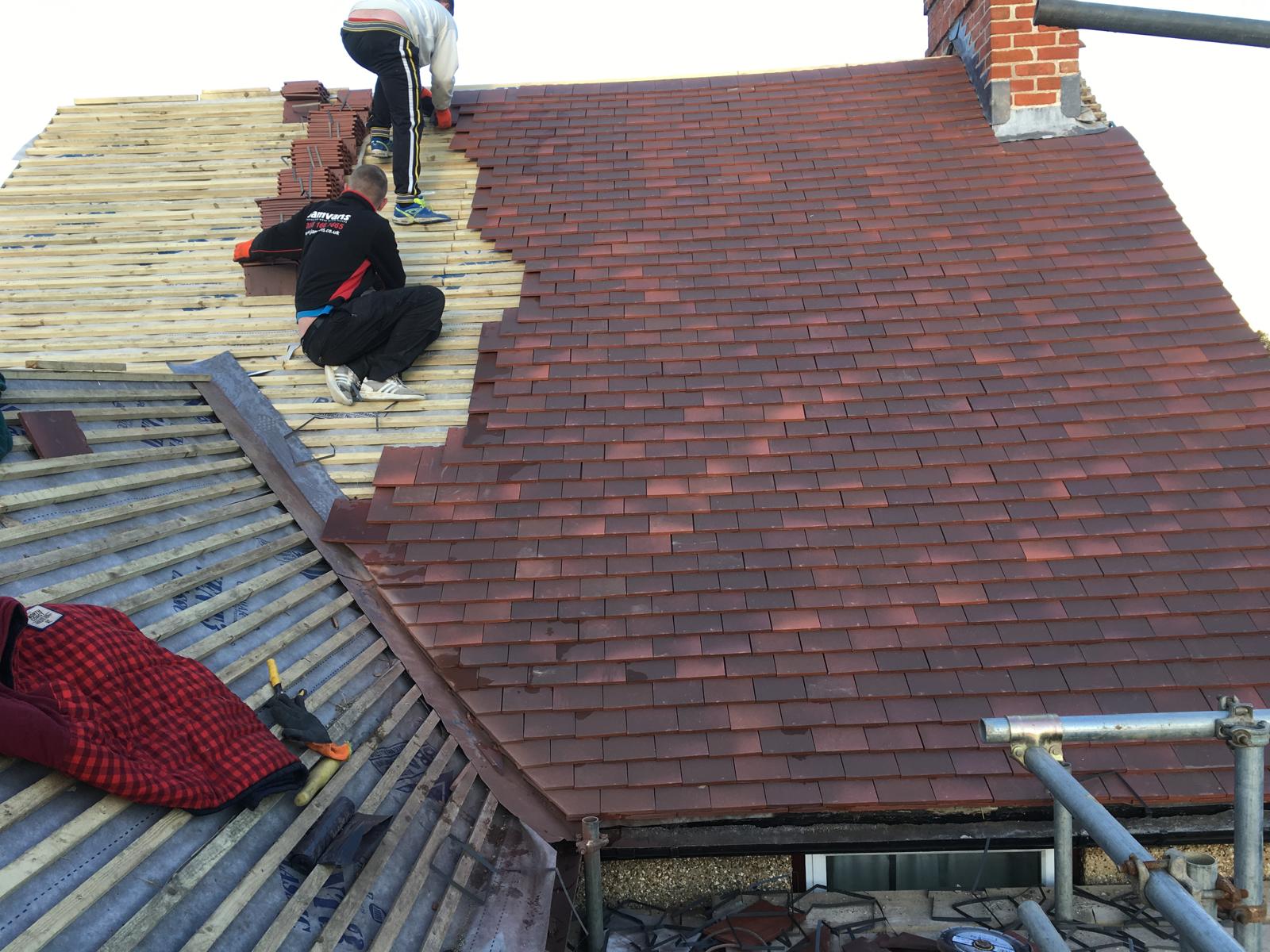 New red clay plain tiles being laid over fresh battens beside a lead valley, two roofers working up the slope